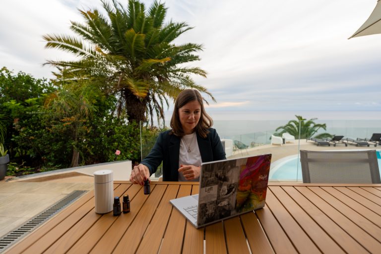 Julia am Terrassentisch mit Laptop Ölen und Diffuser im Hintergrund Palme